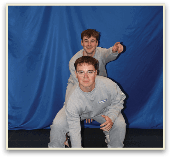 Two young men are posing for a picture, one kneeling and the other standing. They are both wearing grey shirts. AI generated content