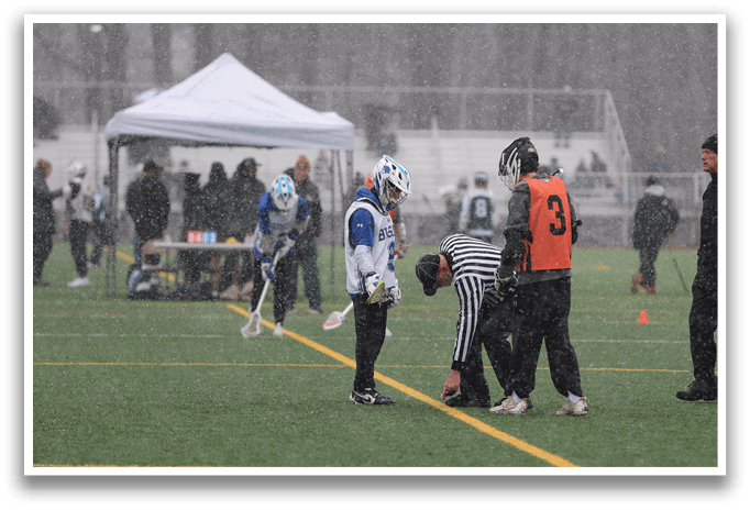 A group of people are standing on a field in the rain, with some of them wearing helmets. One person is kneeling down, possibly injured. The scene appears to be a lacrosse game, as there are lacrosse sticks visible. AI generated content