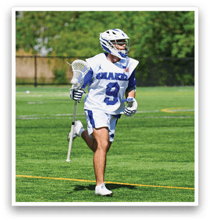 A man wearing a white and blue shirt and shorts is holding a lacrosse stick. Description generated by AI