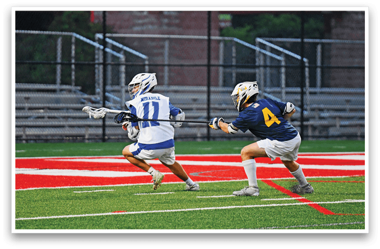 Two people wearing helmets and sports uniforms are playing lacrosse on a field with red and white markings. Description generated by AI