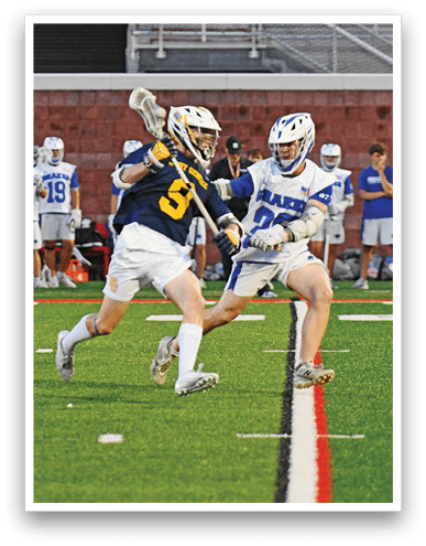 A person wearing a navy blue sports jersey with the number 5 and white shorts is running on a green field while holding a lacrosse stick, with other players and a brick wall in the background. Description generated by AI