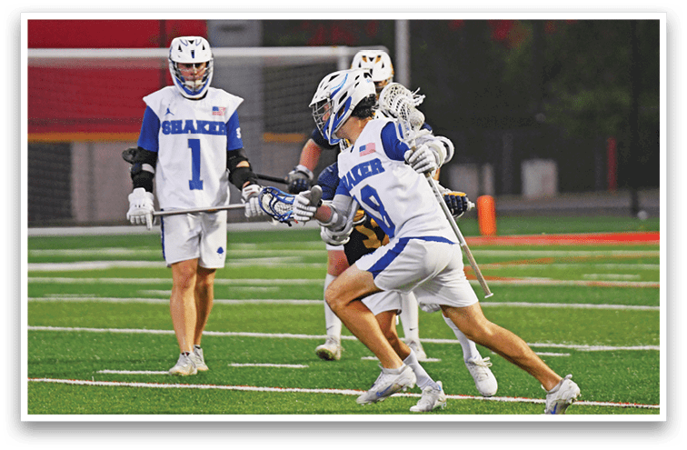 Three people wearing lacrosse gear are playing on a field with one person in a white and blue uniform holding a lacrosse stick and crouching while two others stand nearby. Description generated by AI