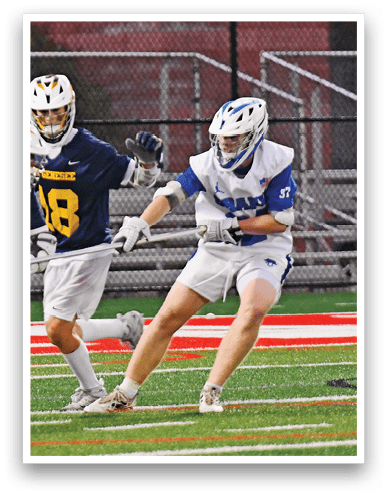 Two people wearing helmets and sports uniforms are playing lacrosse on a field with a red and white sideline. Description generated by AI