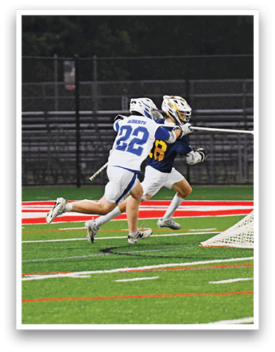 Two people wearing helmets and sports uniforms are playing lacrosse on a field near the goal. Description generated by AI