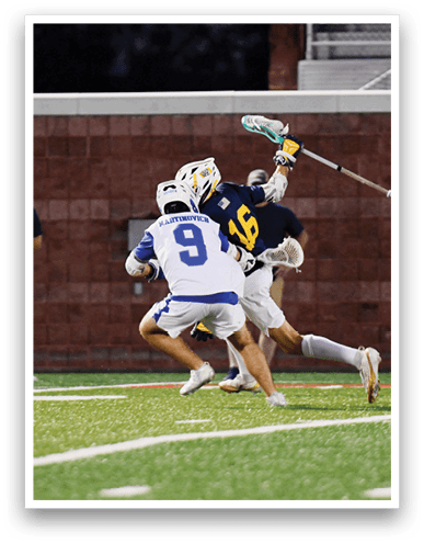 Two people in sports uniforms and helmets are actively playing lacrosse on a field while a person in casual clothing stands in the background. Description generated by AI