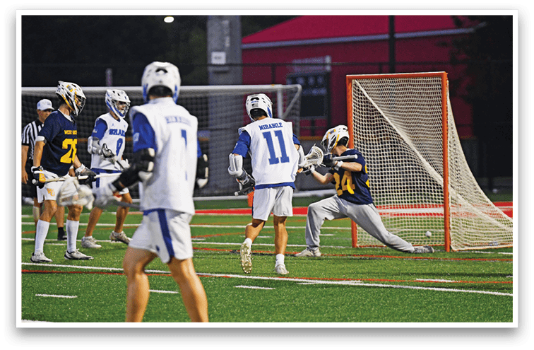 People wearing helmets and sports uniforms are playing lacrosse on a field with one person in a dark uniform attempting to block a shot near the goal. Description generated by AI