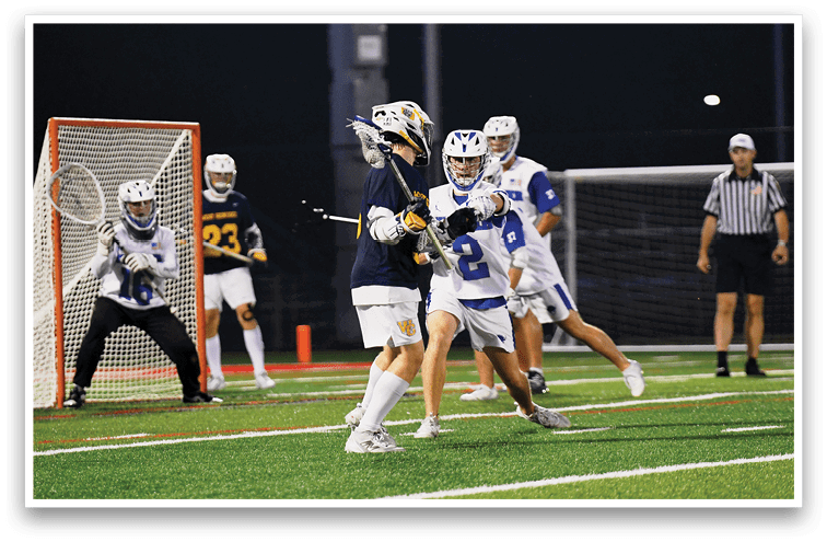 A person in sports gear is positioned as a goalie in front of a lacrosse goal while two other players stand nearby on a field at night. Description generated by AI