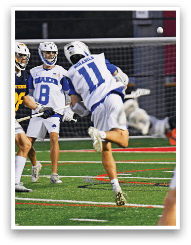 People wearing helmets and sports uniforms are actively playing lacrosse on a field with a goal net in the background. Description generated by AI