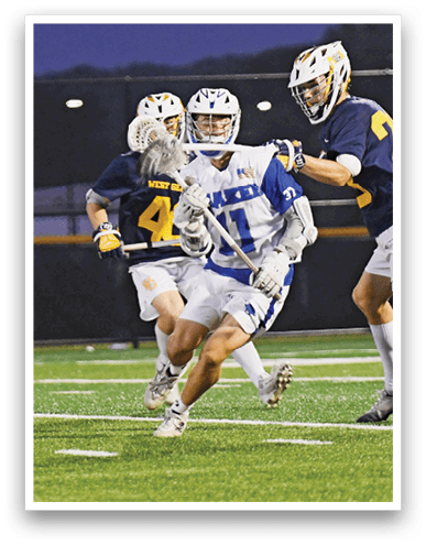 A person wearing a helmet and sports uniform is running on a field while holding a lacrosse stick during a nighttime game. Description generated by AI