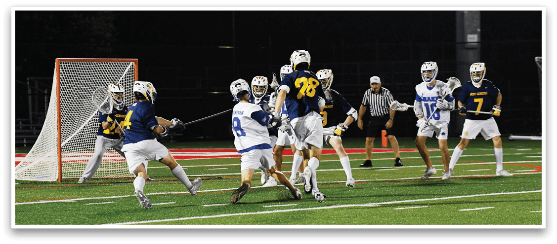 People in sports uniforms are playing lacrosse on a field at night with one person preparing to shoot the ball toward the goal while others defend and a referee watches. Description generated by AI