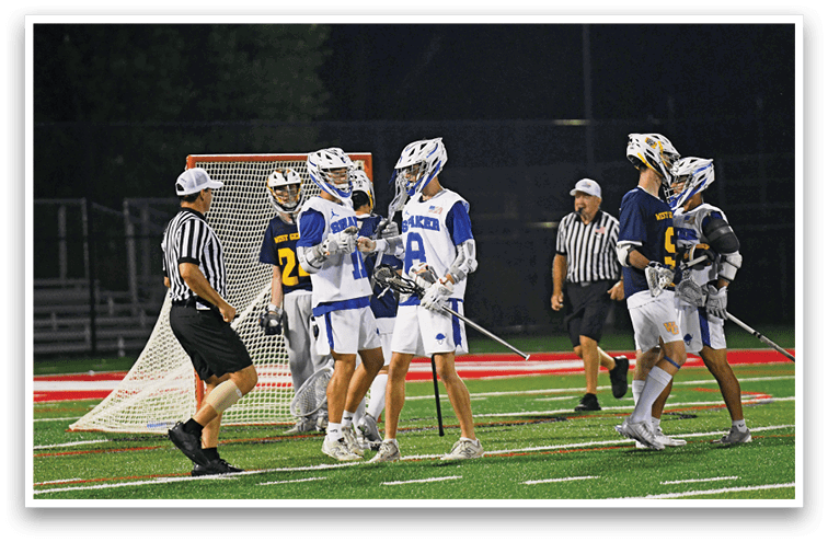 Players in lacrosse uniforms and helmets are gathered on a field near a goal with two referees in striped shirts observing the scene. Description generated by AI
