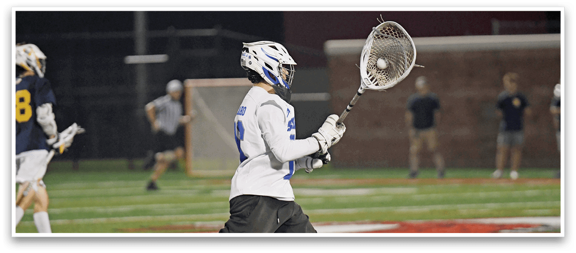 A person in white sports gear and helmet is holding a lacrosse stick with a ball in the netting on a field at night with other players and a referee in the background. Description generated by AI