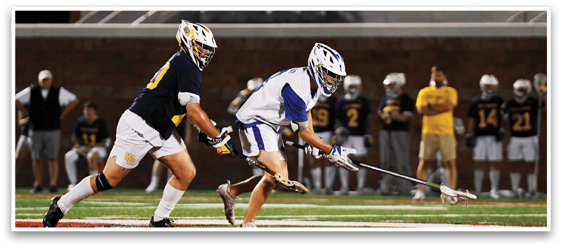 Two people in sports uniforms and helmets are actively playing lacrosse on a field with others watching in the background. Description generated by AI