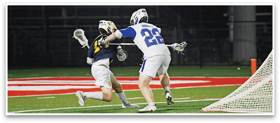Two people wearing helmets and sports uniforms are playing lacrosse on a field near a goal. Description generated by AI
