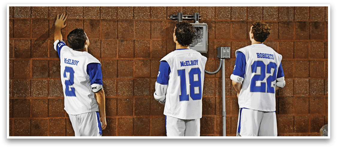 Three people wearing sports jerseys with numbers and names on the back stand facing a brick wall, with one person reaching up to touch the wall. Description generated by AI