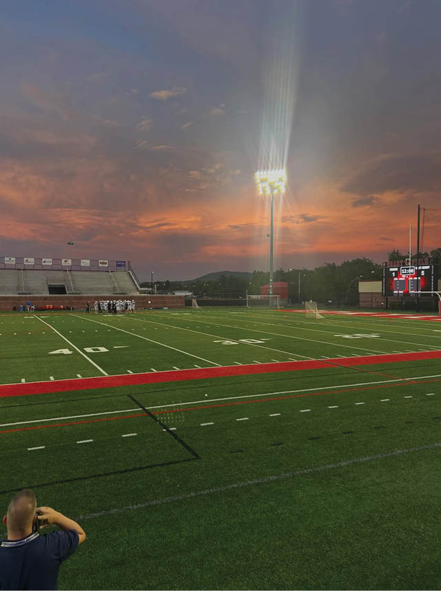 A person stands on the edge of a football field at sunset with stadium lights illuminating the field and a scoreboard visible in the background. Description generated by AI