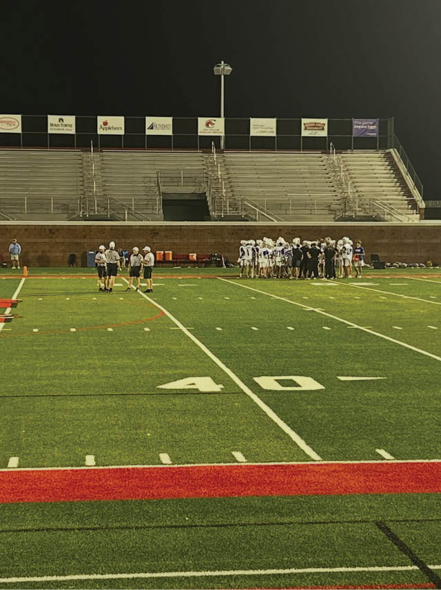 A group of people in sports uniforms are gathered on a football field near the 40-yard line under stadium lights with empty bleachers in the background at night. Description generated by AI