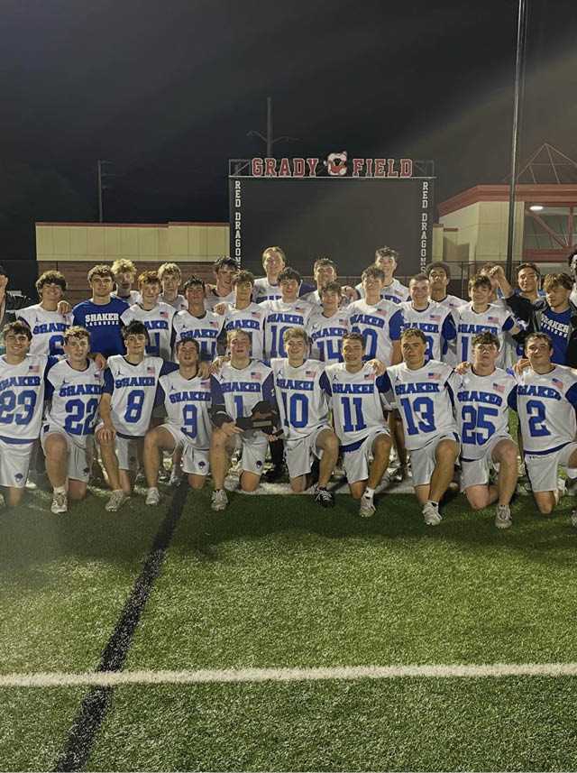A group of people wearing white and blue sports uniforms pose together on a field at night in front of a scoreboard that reads Grady Field. Description generated by AI