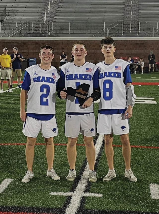 Three people wearing white and blue Shaker sports uniforms stand together on a football field at night with empty bleachers in the background. Description generated by AI