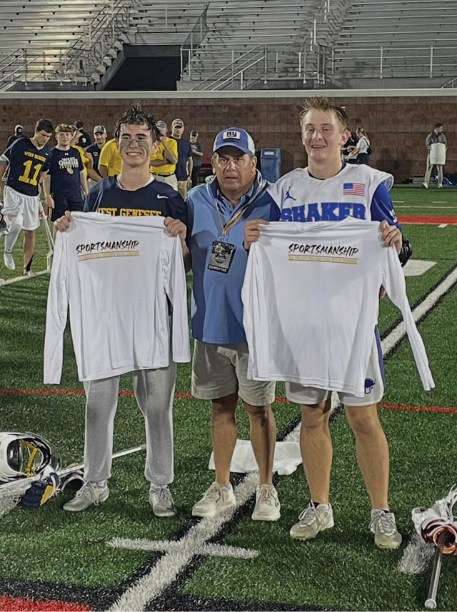 Three people standing on a sports field holding white long-sleeve shirts with the word “SPORTSMANSHIP" printed on them, with one person in the middle wearing a blue jacket and cap and the other two wearing athletic clothing. Description generated by AI