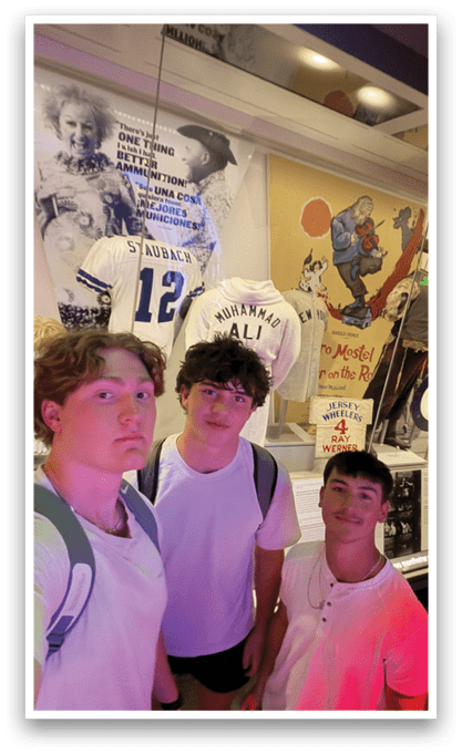 Three young men pose for a picture in front of a museum display featuring sports jerseys and other memorabilia. AI generated content