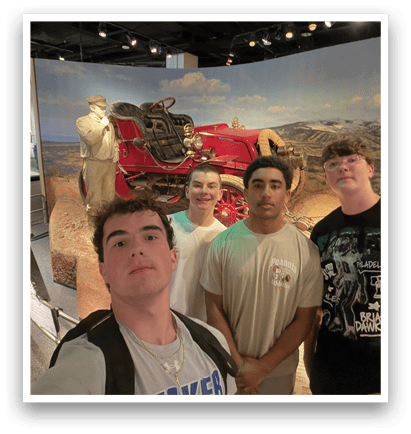 Four young men pose for a picture in front of a display featuring an old-fashioned car and a man. AI generated content