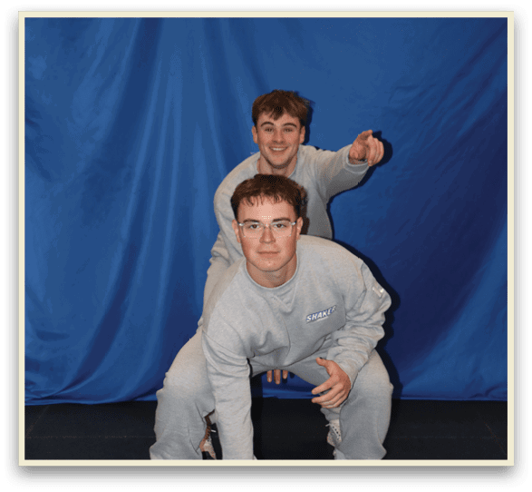 Two young men are posing for a picture, one kneeling and the other standing. They are both wearing grey shirts. AI generated content