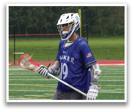 A man wearing a blue shirt and grey pants holding a lacrosse stick. Description generated by AI