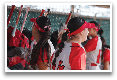 A group of young women are standing in a dugout, wearing red and white uniforms. They are holding baseball bats and are likely waiting for their turn to play. AI generated content