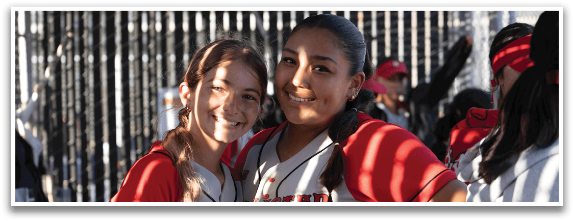 Two girls wearing red and white baseball uniforms. Description generated by AI