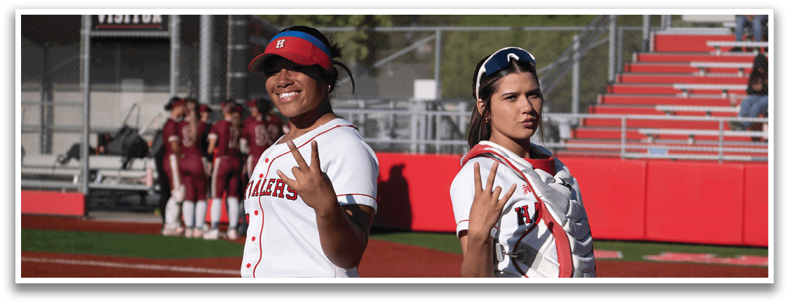 Two women wearing red and white uniforms are standing on a baseball field. Description generated by AI