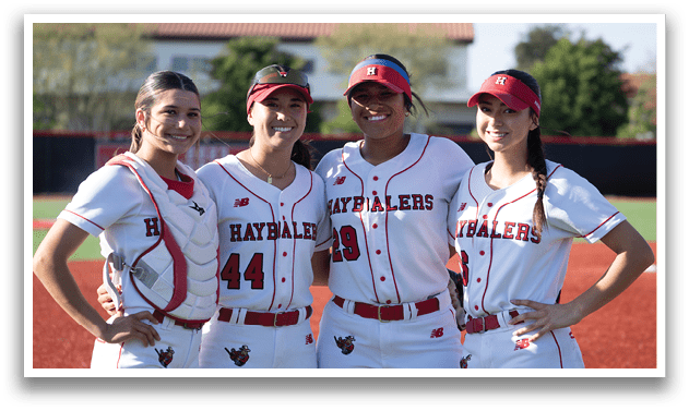 Four women wearing baseball uniforms and hats. Description generated by AI