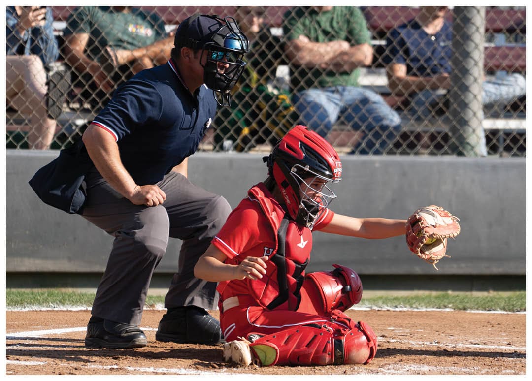 A baseball game is in progress with a batter, catcher, and umpire on the field. The batter is holding a bat, ready to swing, while the catcher is wearing a baseball glove. The umpire is standing behind the catcher, closely observing the play. There are several other people in the background, likely spectators or teammates. AI generated content