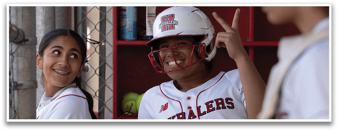 Two female baseball players are standing next to a fence. One player is wearing a helmet and pointing at the other player. AI generated content