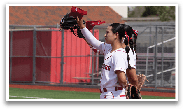 A girl wearing a red hat and white uniform is standing on a baseball field. She is holding a baseball glove and appears to be looking up, possibly at the sky or a ball. AI generated content