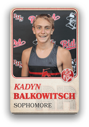 A person wearing a sleeveless sports jersey with a baseball player graphic stands in front of a backdrop featuring the letters RHS and the word Riders, with the name Kadyn Balkowitsch and the word Sophomore displayed below. Description generated by AI