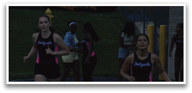 Two women are running on a track, one wearing a black and white outfit and the other wearing a black and white and black outfit. They are both holding pink and blue glow sticks. Contenido generado con IA