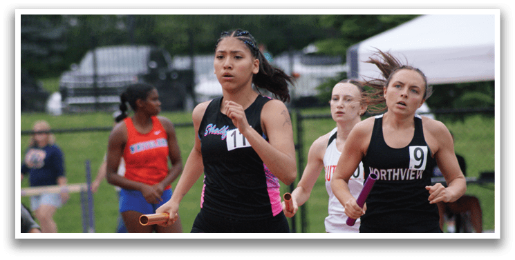 Three female runners are racing on a track, with one wearing a green shirt and the other two wearing red. Contenido generado con IA