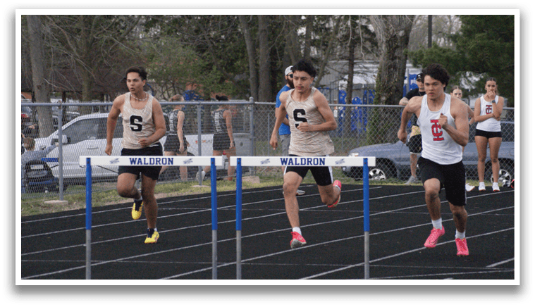 A group of people are running on a track, with some of them wearing numbers on their shirts. They are running in front of a blue and white barrier. Contenido generado con IA