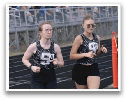 Two girls are running on a track, one wearing a black shirt and the other wearing a white shirt. They are both wearing sunglasses. Contenido generado con IA