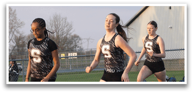 Three female runners are on a track, wearing black and white uniforms. They are running in a line, with one in the lead and the other two following closely behind. Contenido generado con IA