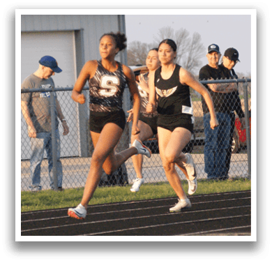 Two girls are running on a track, with one girl leading the other. They are wearing shorts and are in the middle of a race. There are several other people watching the race from the sidelines. Contenido generado con IA