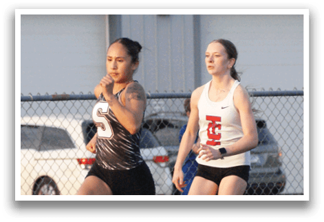 Two girls are running on a track, one wearing a black shirt and the other wearing a white shirt. They are both wearing shorts. Contenido generado con IA