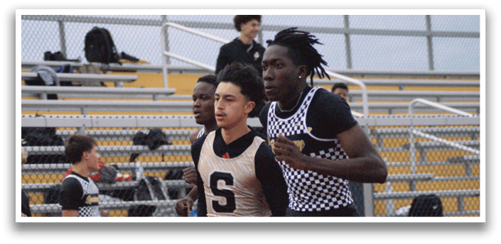 Two young men are running on a track, one wearing a black shirt and the other wearing a white shirt. They are both running towards a barrier. Contenido generado con IA