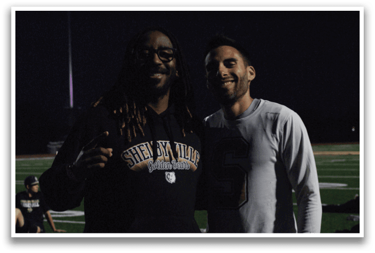 Two men are standing on a field, one wearing a hoodie with the word “Shea" on it. Contenido generado con IA