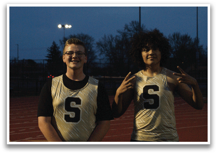 Two young men wearing sports jerseys pose for a picture on a track. Contenido generado con IA
