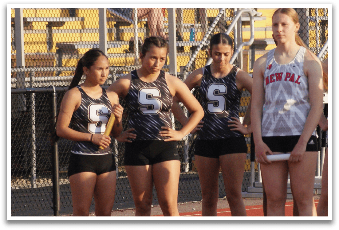 A group of female athletes are standing on a track, wearing uniforms and holding their hands together. They are lined up in front of a crowd of people who are watching them. Contenido generado con IA