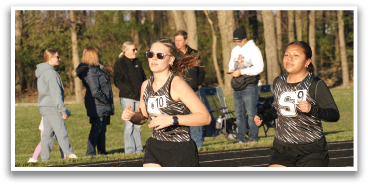 Two female runners are running on a track, with one wearing a black and white outfit and the other wearing a black and white and gray outfit. They are running in front of a crowd of people who are watching them. Contenido generado con IA