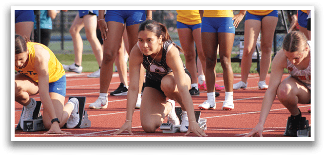 A group of female athletes are lined up on a track, wearing shorts and shirts. They are in various positions, some crouching and others standing. Contenido generado con IA