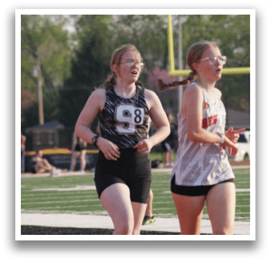Two girls are running on a field, one wearing a white shirt and the other wearing a black shirt. They are both holding water bottles. Contenido generado con IA